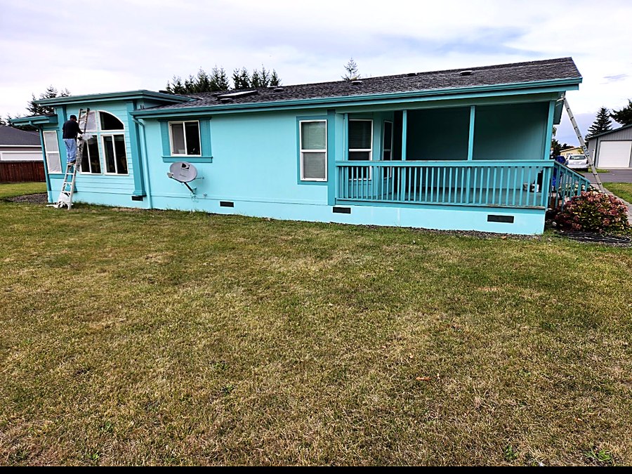Single-story home repainted in bright turquoise with white window trim and a matching porch rail, a painter on a ladder finishing the far gable.